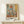 May include: Framed abstract art print in warm wood frame displaying geometric shapes in muted blues, oranges, and earth tones, leaning against white wall on light wooden surface beside small white wrapped gift with natural twine and evergreen sprig