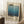 May include: A framed Mark Rothko exhibition poster featuring a blue gradient watercolor wash transitioning from light to deep blue, with black text displaying "Mark Rothko" and exhibition dates "02.19.1970". The poster is displayed in a light wood frame, leaning against white wainscoted walls on a white textured rug. A coffee cup sits on a wooden side table nearby, with soft natural lighting creating shadows across the scene.