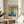 May include: Framed abstract art print with blue vertical band intersecting orange and yellow horizontal sections, displayed on round wooden side table next to beige upholstered chair. Natural sunlight creates geometric shadows on white wall. Green plant leaves visible in corner. Print features French text at top and artist name below artwork in thin golden frame.