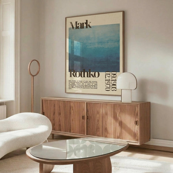 May include: Vintage-style Mark Rothko exhibition poster with blue geometric artwork displayed above wooden sideboard in modern living room. Poster shows cream background with blue rectangular forms and black text including "Rothko" and dates. Room features white curved chair, glass oval coffee table, and warm wood furniture against neutral walls.