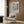 May include: Beige upholstered armchair with geometric patterned pillow positioned against wood-paneled wall, framed vintage art poster featuring blue abstract squares and text mounted above chair, brass floor lamp with white circular base beside chair, dark round side table with open book, neutral curtains by window, light wood flooring