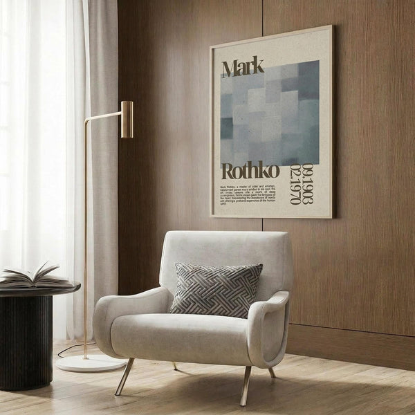 May include: Beige upholstered armchair with geometric patterned pillow positioned against wood-paneled wall, framed vintage art poster featuring blue abstract squares and text mounted above chair, brass floor lamp with white circular base beside chair, dark round side table with open book, neutral curtains by window, light wood flooring