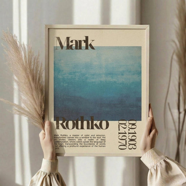 May include: Person holding framed poster featuring blue gradient artwork with "Mark Rothko" text and biographical description. Light wooden frame against cream background with dried pampas grass visible. Person wears beige ruffled sleeve top. Poster shows ocean-like blue tones transitioning from light to dark with vintage typography and exhibition-style layout.