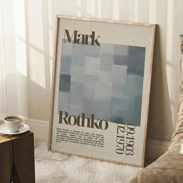 May include: A framed Mark Rothko exhibition poster leaning against a white wall in a cozy room setting. The poster features abstract blue and gray rectangular color blocks with "Mark Rothko" text and exhibition details "04.17-08.1.60". A white coffee cup sits on a wooden surface nearby, with a beige knit blanket and white textured rug visible in the soft natural lighting.