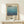 May include: Framed poster featuring Mark Rothko's name and exhibition details with a blue gradient square artwork transitioning from light blue at top to deep navy at bottom. The minimalist print has vintage exhibition text and dates. Shown in light wood frame on white wooden dresser next to small wrapped gift with twine bow against cream wall.