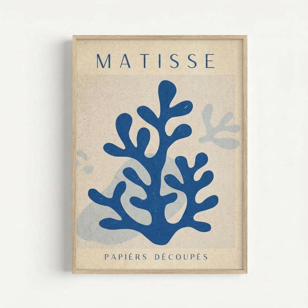 Framed poster featuring blue coral-like cut-out shapes in Matisse's paper collage style on cream textured background. "MATISSE" appears at top in dark blue letters, "PAPIERS DÉCOUPÉS" at bottom. Light wood frame against white wall. Main coral form is deep blue with organic branching shapes, lighter blue-gray forms visible in background.