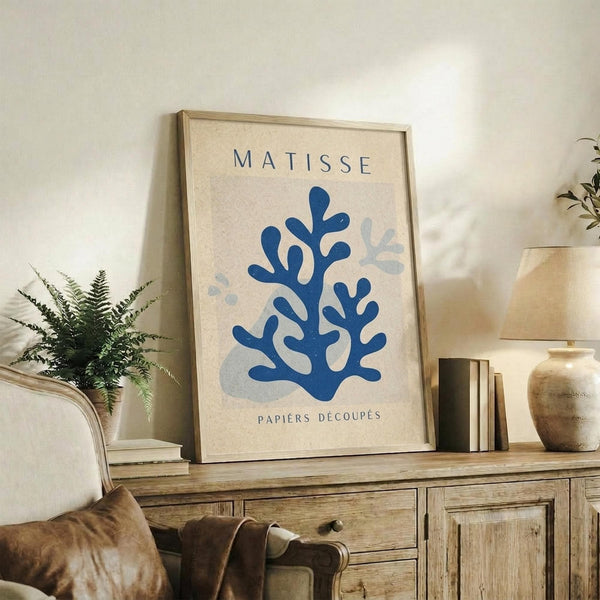 May include: Framed beige poster with blue coral-like cut-out shapes in Matisse style, displayed on rustic wooden sideboard alongside fern plant, ceramic vase, and books in neutral-toned living room with cream armchair and brown throw pillows