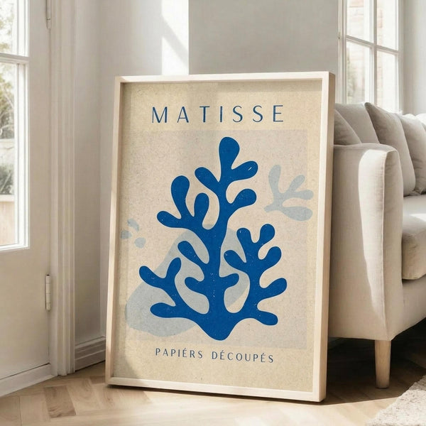May include: Framed poster with beige background featuring blue coral-like cut-out shapes in Matisse style, with "MATISSE" and "PAPIERS DÉCOUPÉS" text. Light wooden frame leaning against white wall next to beige upholstered chair in bright, naturally-lit room with white window frames and hardwood flooring.