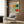 May include: A cream-colored exhibition poster featuring Mark Rothko's name and colorful abstract rectangular blocks in orange, teal, mint green, and coral red, displayed in a light wood frame on a walnut-paneled wall above a beige upholstered armchair with geometric throw pillow, beside a brass floor lamp and black side table in a modern living room setting.