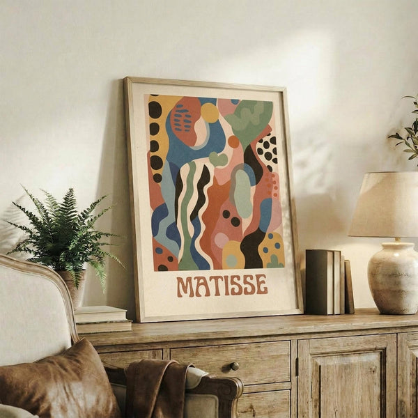 May include: Colorful abstract art print with organic shapes in blues, oranges, greens, and coral tones, featuring flowing lines and dotted patterns in the style of mid-century modern art. Framed in light wood and displayed on a rustic wooden sideboard alongside a fern plant, ceramic vase, and books in a cozy living room setting with neutral walls and warm lighting.