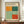 May include: A framed Mark Rothko exhibition poster displayed on a wooden sideboard in an elegant room with white paneled walls. The poster features abstract geometric color blocks in orange, teal, sage green, and coral red on a cream background. A small white ceramic vase and stack of books sit beside the brown-framed artwork, which shows exhibition details and dates.