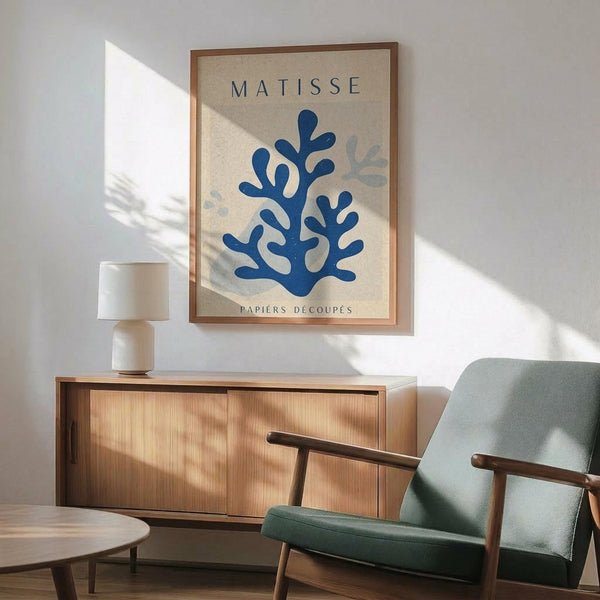 May include: Modern living room with mid-century wooden sideboard, sage green armchair, and round coffee table. Framed art print on white wall features blue coral-like cutout shapes on cream background with "MATISSE" and "PAPIERS DÉCOUPÉS" text. Natural sunlight creates dramatic shadows across the scene. White ceramic table lamp sits on sideboard.