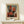 May include: Abstract painting poster with overlapping geometric rectangles in red, orange, black and gray tones on muted background, displayed in thin wooden frame on light wood surface beside small white wrapped gift with evergreen sprig against cream wall