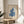 May include: Framed poster with Matisse-inspired blue coral or seaweed cut-out design on cream background, displayed on round wooden side table next to beige upholstered chair. Natural sunlight streams across white walls creating geometric shadows. Green plant leaves visible in corner of minimalist, neutral-toned room setting.