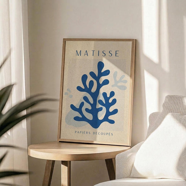 May include: Framed poster with Matisse-inspired blue coral or seaweed cut-out design on cream background, displayed on round wooden side table next to beige upholstered chair. Natural sunlight streams across white walls creating geometric shadows. Green plant leaves visible in corner of minimalist, neutral-toned room setting.