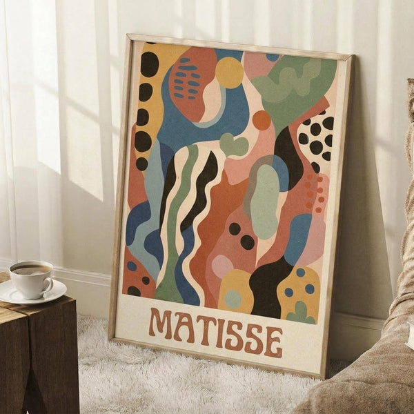 May include: A large framed poster with colorful abstract shapes in blues, oranges, greens, and yellows featuring organic forms, dots, and wavy lines in a Matisse-inspired style. The artwork leans against white paneled walls beside a wooden side table with coffee cup and blanket, displayed on a textured rug in a cozy room setting.
