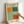 May include: Two hands holding a framed exhibition poster featuring abstract color blocks in orange, teal, mint green, and coral red against a cream background. The poster displays Mark Rothko gallery text with exhibition dates. Light wood frame against white wall with soft natural lighting creating gentle shadows.