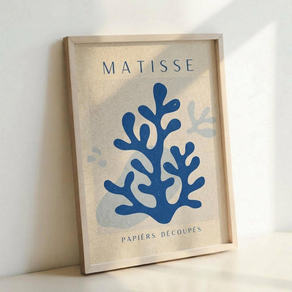 May include: Framed art print in light wood frame leaning against white wall. Poster features blue coral-like cut-out shapes on cream textured background with "MATISSE" at top and "PAPIERS DÉCOUPÉS" at bottom in navy text. Additional lighter blue organic shapes scattered around main design. Natural lighting creates soft shadows on wall.