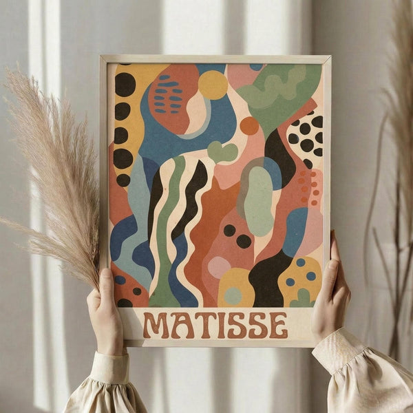 May include: Person holding framed abstract art print in natural wood frame against white curtained window. Colorful composition features organic shapes in coral pink, sage green, navy blue, cream, and golden yellow with black dots and wavy lines. Dried pampas grass visible on left side. Print displays "MATISSE" text at bottom in brown lettering.