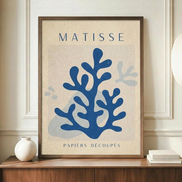May include: Framed art print featuring blue coral-like cutout shapes in Matisse's papier découpé style on cream textured background, displayed on wooden sideboard next to white ceramic vase and stacked books in elegant room with white wainscoting and warm lighting.