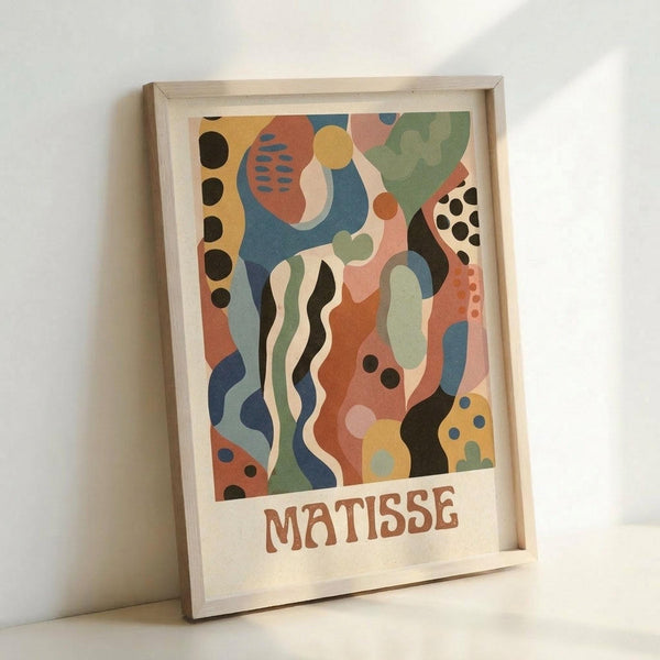 May include: Framed abstract art print in light wood frame leaning against white wall. Artwork features flowing organic shapes in coral pink, sage green, navy blue, mustard yellow, and cream with scattered dots and wavy lines. "MATISSE" text appears at bottom in brown letters. Modern minimalist styling with natural lighting casting soft shadows.
