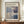 May include: Framed Mark Rothko exhibition poster featuring abstract rectangular color blocks in muted blues, purples, beige and cream tones. The poster displays gallery information and April-July 2023 dates. Shown in thin brown frame on wooden sideboard alongside white ceramic vase and stacked books against cream paneled wall.