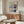 May include: Modern living room with Mark Rothko exhibition poster featuring abstract color blocks in blues, grays, and cream tones. Poster hangs above mid-century walnut sideboard with brass circle lamp. White curved chair and glass oval coffee table complete the minimalist Scandinavian-style interior with natural lighting.