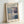May include: Framed exhibition poster featuring Mark Rothko's abstract color field painting with muted blue, beige, and purple rectangular blocks. Light wood frame displays cream-colored poster with black text about Rothko Art Gallery NYC exhibition dates April-July 2023. Frame leans against white wall on light surface with soft natural lighting creating gentle shadows.