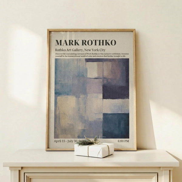 May include: Abstract art exhibition poster in light wood frame leaning against white wall on wooden shelf. Features geometric color blocks in blues, grays, and cream tones with black text header. Small white wrapped gift with greenery sits in front. Minimalist Scandinavian-style home decor setting with natural lighting and neutral color palette.