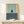 May include: Abstract minimalist artwork in wooden frame displayed on light wood surface. Composition features pale green upper section with small dark blue rectangle and thin vertical line, cream colored side panel, and darker blue-gray lower section with white text. Small wrapped gift with evergreen sprig sits nearby against neutral wall background.