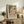 May include: Framed poster with colorful abstract artwork featuring green foliage, yellow citrus fruits, dark tree trunks, and pink ground in Matisse's distinctive style. Black text below describes the exhibition. Displayed on rustic wooden sideboard alongside fern plant, books, and ceramic vases in neutral-toned living room with beige upholstered furniture.