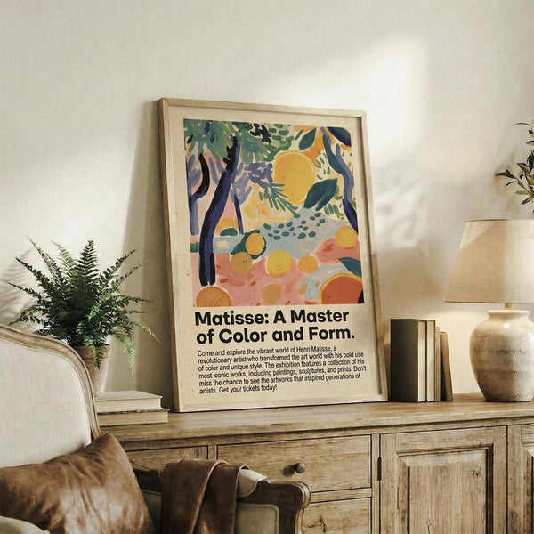 May include: Framed poster with colorful abstract artwork featuring green foliage, yellow citrus fruits, dark tree trunks, and pink ground in Matisse's distinctive style. Black text below describes the exhibition. Displayed on rustic wooden sideboard alongside fern plant, books, and ceramic vases in neutral-toned living room with beige upholstered furniture.
