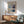 May include: Mark Rothko exhibition poster in thin wooden frame hanging on white wall above light wood credenza with table lamp. Mid-century modern sage green armchair with wooden arms sits nearby. Poster features abstract rectangular color blocks in blues, yellows, and grays with gallery text and exhibition dates. Natural sunlight creates geometric shadows across the minimalist room scene.