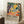 May include: Framed poster with vibrant Matisse-inspired artwork featuring orange circles, green foliage, dark tree trunks, and coral-pink ground in fauve style. Black exhibition text below reads "Matisse: A Master of Color and Form" with description. Light wood frame leaning against white wall corner beside beige armchair and coffee cup on wooden side table.