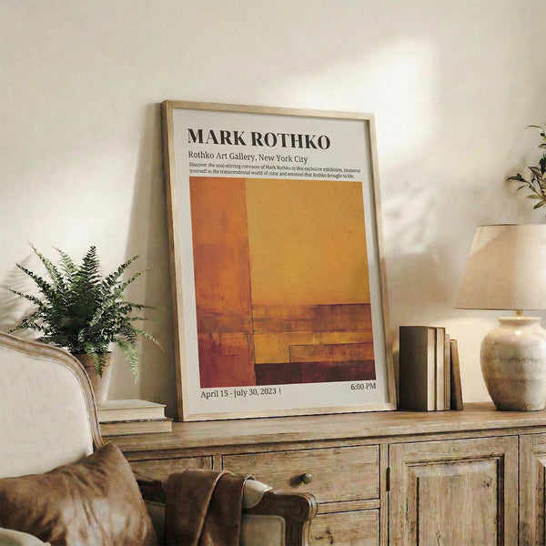 May include: Framed exhibition poster featuring abstract color blocks in warm orange, yellow, and brown tones displayed on a rustic wooden console table alongside books, potted fern, ceramic vase, and beige upholstered chair in a neutral-toned living room setting.