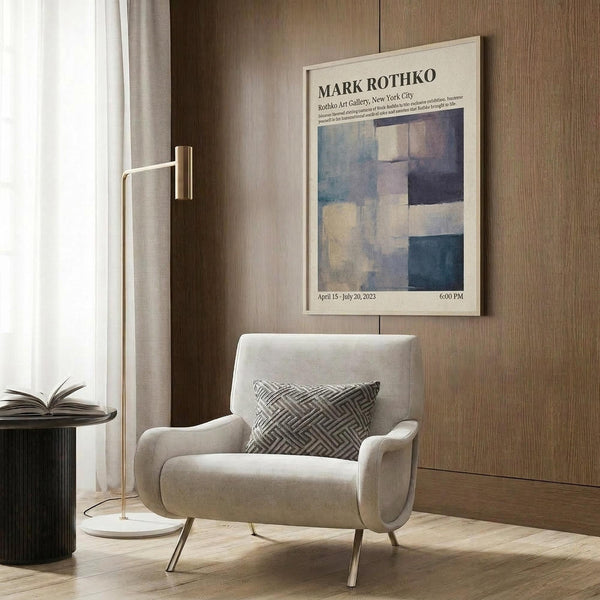 May include: Light beige armchair with geometric patterned pillow positioned near wood-paneled wall. Framed art poster featuring abstract blue and beige color blocks hangs above. Modern brass floor lamp with white circular base stands nearby next to dark round side table with open book. Warm natural lighting from window with sheer curtains creates cozy reading nook atmosphere.