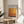 May include: Framed exhibition poster featuring Mark Rothko artwork with warm orange, yellow, and burgundy color blocks displayed on a round wooden side table next to a beige upholstered chair. Natural sunlight streams through white curtains, casting soft shadows across the minimalist room setting. Green plant leaves visible in corner.