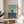 May include: Abstract art print in thin gold frame displayed on round wooden side table next to beige upholstered chair. Artwork features geometric composition with light blue-green upper section, yellow vertical stripe on left, small dark blue rectangle in center, and darker blue-gray lower portion. Natural sunlight creates soft shadows across white wall and neutral home decor setting.