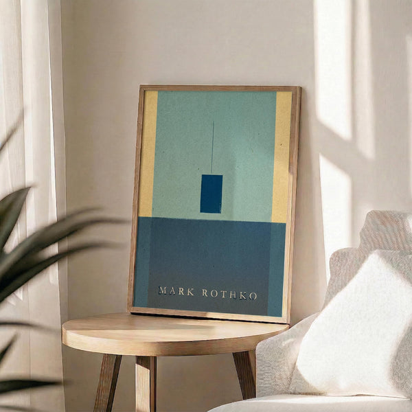 May include: Abstract art print in thin gold frame displayed on round wooden side table next to beige upholstered chair. Artwork features geometric composition with light blue-green upper section, yellow vertical stripe on left, small dark blue rectangle in center, and darker blue-gray lower portion. Natural sunlight creates soft shadows across white wall and neutral home decor setting.