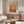 a modern living room with a large abstract painting hanging on the wall above a wooden cabinet. The painting has a warm color palette and depicts a landscape scene. The room also includes a white sofa, a glass coffee table, and a lamp on a side table.