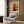 May include: Abstract art poster with warm orange, yellow and brown color blocks in white frame, hung on wood paneled wall above beige armchair with geometric pillow, brass floor lamp and side table with open book nearby