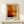 May include: Framed abstract art print in warm orange, yellow, and cream color blocks with dark horizontal lines, displayed on light wood console table beside small white wrapped gift with greenery sprig against cream wall