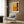 May include: Modern living room with beige armchair and geometric pillow, brass floor lamp, dark round side table with open book, wood paneled wall. Framed abstract art poster featuring warm orange, yellow, and burgundy color blocks in minimalist style hangs above chair. Natural lighting from window with white curtains.