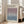 May include: Framed art poster leaning against white wall next to beige sofa in bright living room. Poster features gradient artwork transitioning from purple at top to blue at bottom, with exhibition text and dates. Light wood frame, natural lighting from windows, hardwood floors visible.