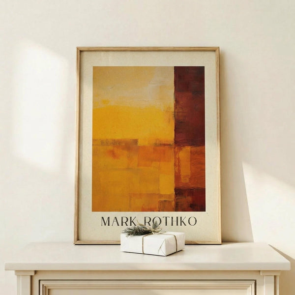 May include: Abstract art print in warm tones displayed in light wood frame on pale wooden surface. Artwork features geometric blocks of yellow, orange, and deep red colors with soft, painterly edges. Small white gift box with natural twine and greenery sits beside the framed piece against cream wall background.