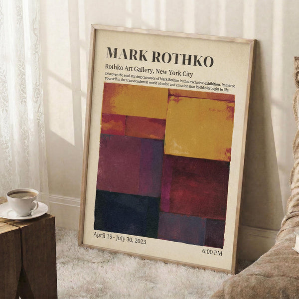 May include: A framed exhibition poster for Mark Rothko's art gallery show in New York City, featuring abstract color blocks in warm yellows, deep reds, purples, and dark blues. The poster displays exhibition dates April 15 - July 30, 2023. The frame leans against a white wall on a textured cream rug, next to a wooden side table with a white coffee cup and saucer. Soft natural light creates shadows across the scene.