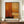 May include: Abstract expressionist artwork with warm orange, yellow, and cream rectangular color blocks separated by dark horizontal lines, displayed in thin brown frame on wooden sideboard. White ceramic vase and stacked books sit nearby. Room features white paneled walls and warm lighting.