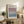 May include: A framed exhibition poster with Mark Rothko's name in bold black text at top, featuring a gradient artwork transitioning from purple at top to blue at bottom. The cream-colored poster shows gallery details and dates April 15 - July 30, 2023. Displayed in a light wood frame on a rustic wooden sideboard alongside books, a ceramic vase, and fern plant, with a beige upholstered chair visible in a neutral-toned living room setting.