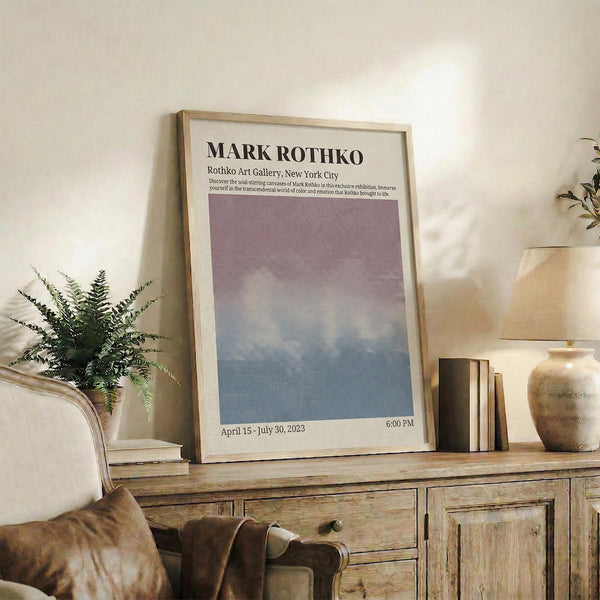 May include: A framed exhibition poster with Mark Rothko's name in bold black text at top, featuring a gradient artwork transitioning from purple at top to blue at bottom. The cream-colored poster shows gallery details and dates April 15 - July 30, 2023. Displayed in a light wood frame on a rustic wooden sideboard alongside books, a ceramic vase, and fern plant, with a beige upholstered chair visible in a neutral-toned living room setting.