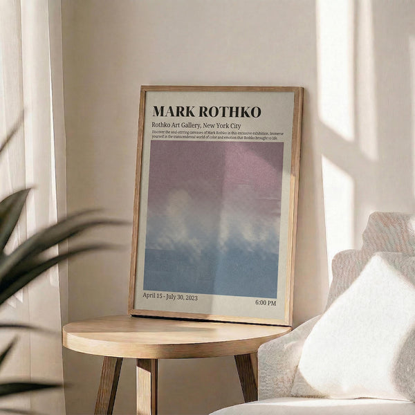May include: Framed exhibition poster with Mark Rothko gallery text and gradient artwork in purple, pink and blue tones, displayed on light wood round side table next to beige upholstered chair with white throw pillow, green plant leaves visible, soft natural window lighting creating shadows on neutral wall